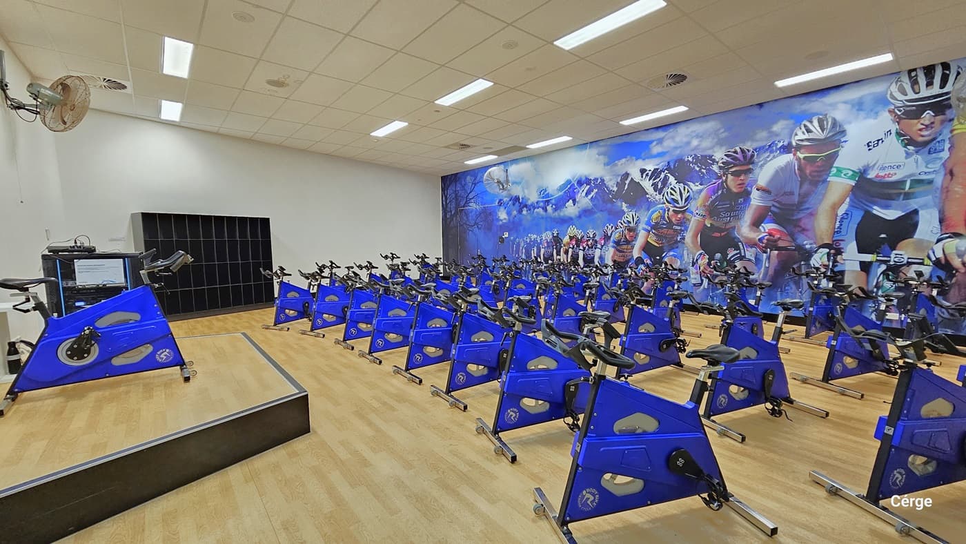 Inside the cycle studio, a row of blue cycle machines is neatly arranged, covering half of the timber floor. On the left, there is an elevated platform with a cycle machine on top, and a fan is mounted on the white wall. An open 4-tier storage cabinet is positioned against the wall. On the right, a large poster, depicting cyclists in motion, covers the entire wall. The area is well-illuminated, with white walls and a white ceiling.