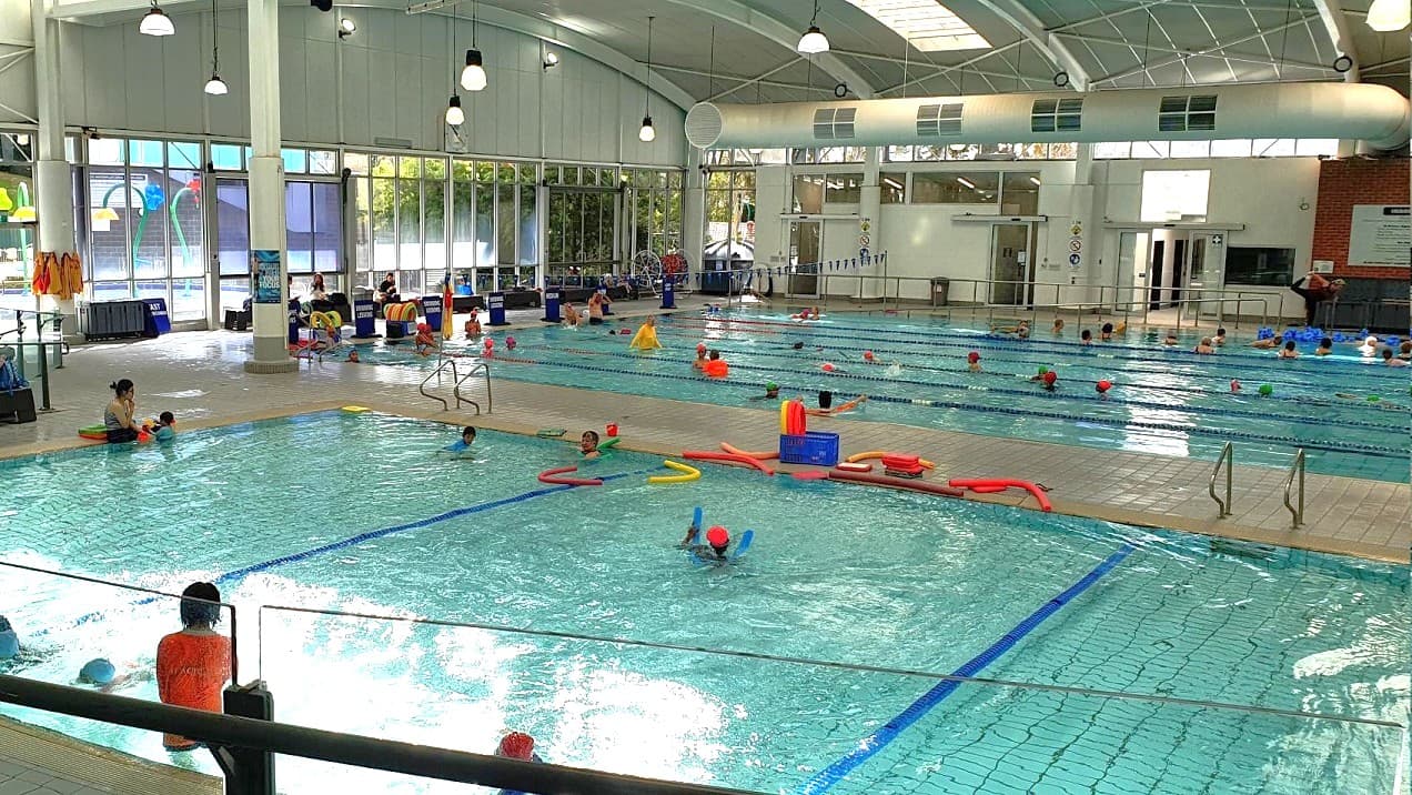 Indoor swimming pool with multiple lanes and people swimming. On the left, a smaller pool area with children and adults, some using floatation devices. The right side has a larger pool with more swimmers. The background features large windows allowing natural light, and a high, arched ceiling with hanging lights. Poolside, there are various swimming aids and equipment. The floor is tiled, and there are steps leading into the water.