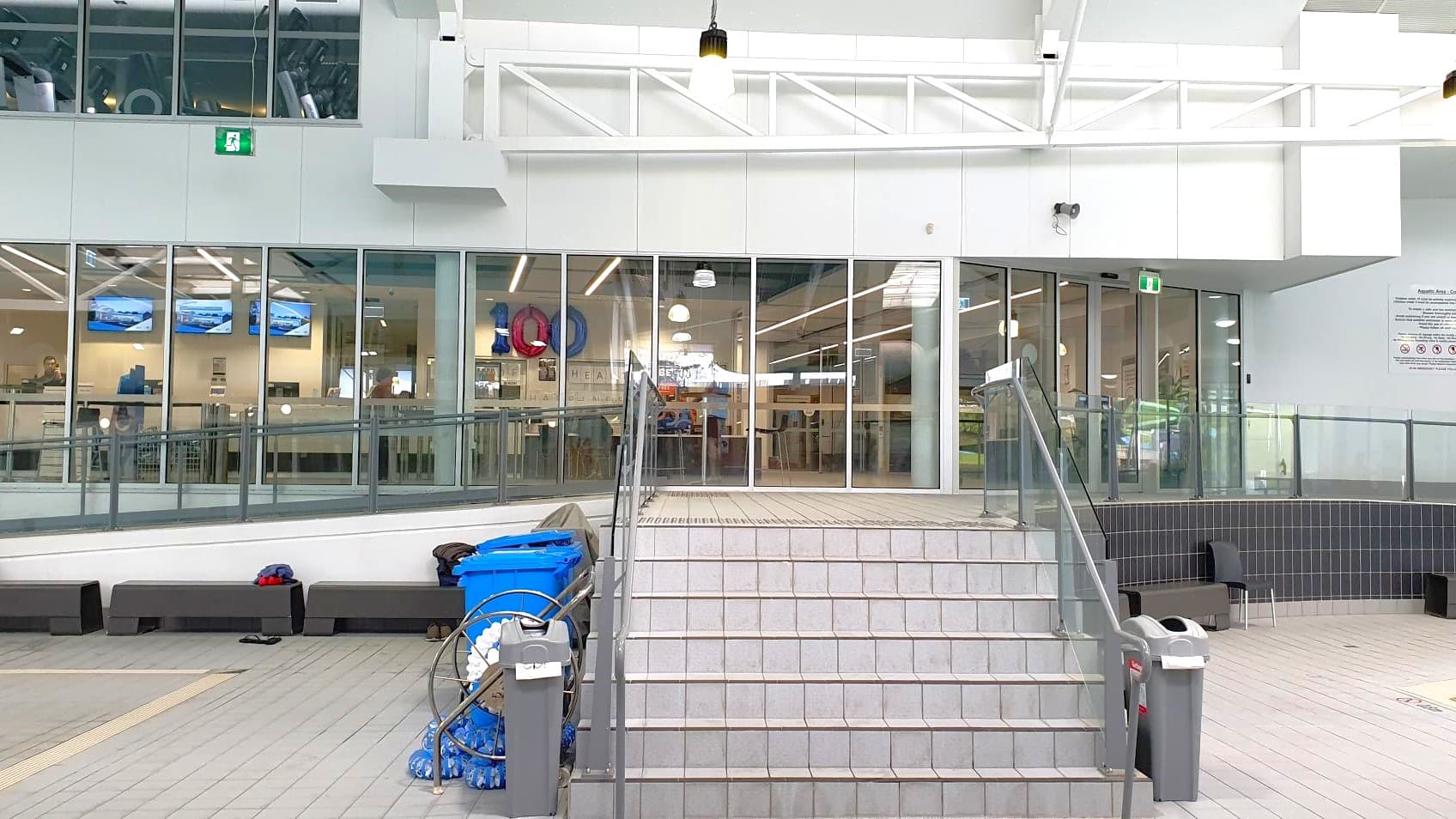 A set of grey tiled stairs leads up to a glass-walled interior space. The glass panels reveal a room with several screens displaying images and a few people inside. On the left, there are blue bins and cleaning equipment. The ceiling is white with exposed beams and hanging lights. The floor is tiled in grey, and there are black benches along the wall. A green exit sign is visible above the glass doors.