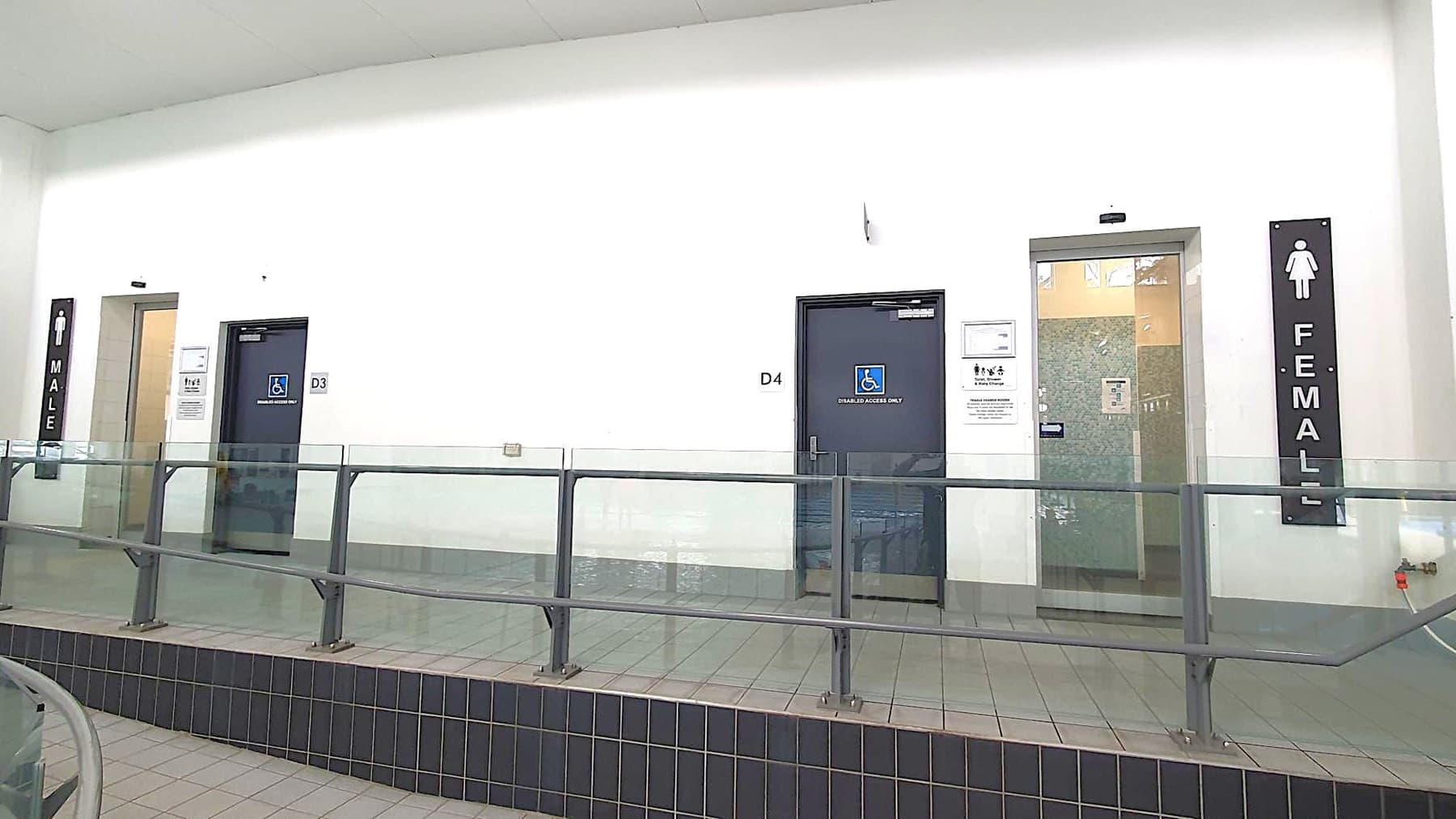 On the left, there is a door marked "MALE" with a sign above it. In the centre, there are two doors, D3 and D4, both with accessibility signs. On the right, a door marked "FEMALE" with a sign above it. A railing runs along the front, and the floor is tiled. The walls are plain white, and the ceiling is not visible.
