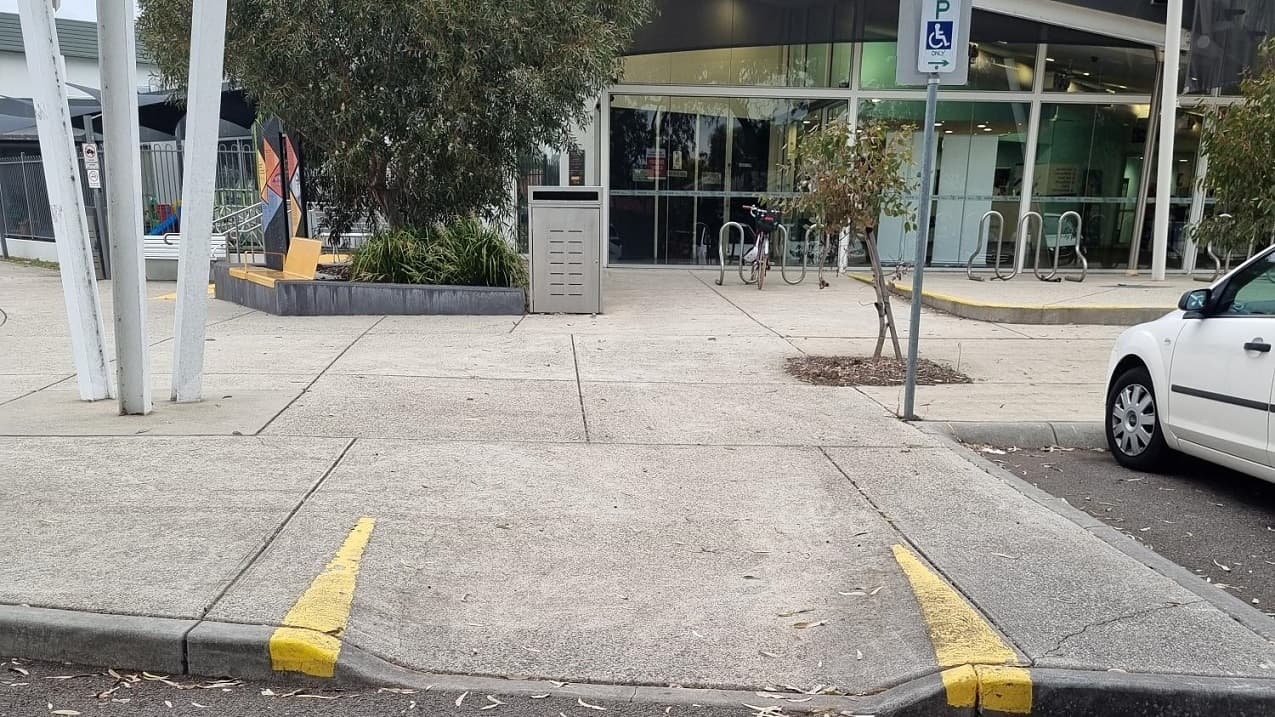 In the foreground, there is a pavement featuring a yellow marking and step-free access. Adjacent to this is a white car parked to the right. Centrally located is a low-set building with a glass facade, in front of which bike racks are positioned. To the left, a letterbox, trees, and banners can be seen. Concrete posts and signage are also visible in the area.