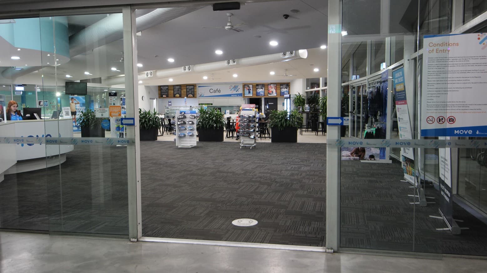 The entrance to the building is via an automatic glass sliding door, which displays a poster detailing entry conditions. Inside, the interior includes a grey carpet and white ceiling equipped with LED lighting and large air conditioning vents. To the left is a reception desk with a white facade, adjacent to a retail display featuring goggles. A large retractable banner stands to the right. In the centre, a large planter box filled with indoor plants is aligned, with a cafe situated behind it.