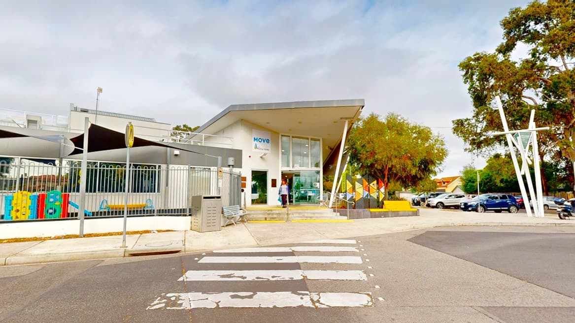 A low-set white building with pitched roofing made of concrete and glass is enclosed by a white metal fence. To the left, there is a planter box with trees and banners. Steps with handrails are adjacent to the building. In the foreground, there is a road featuring a pedestrian lane marked in white. Nearby, a car park is filled with multiple vehicles in various colours. A bench chair and a mailbox are also positioned in front of the building.