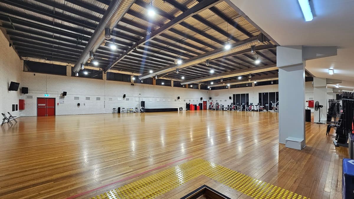 A spacious indoor gym features polished timber flooring and a ceiling decorated with exposed steel beams, large ceiling fans, silver insulation, and strip lighting. Chairs and various pieces of exercise equipment are arranged along the walls, which are made of white bricks. In the left corner, there is a two-tier set of steps fitted with truncated domes.