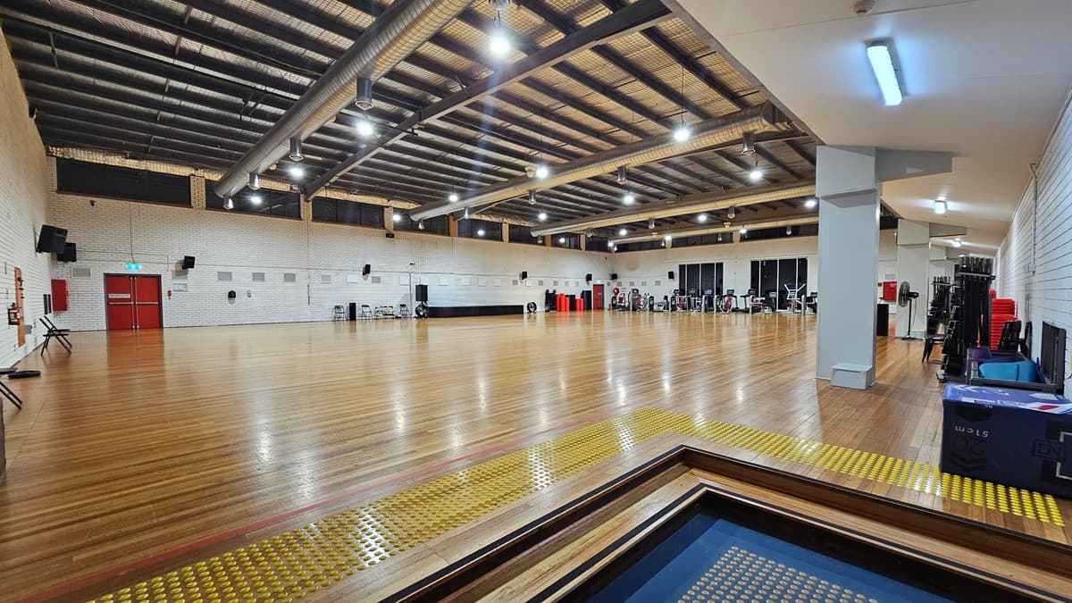A spacious indoor gym boasts polished timber flooring and a ceiling adorned with exposed steel beams, large ceiling fans, silver insulation, and strip lighting. Chairs and various exercise equipment line the walls. The walls are constructed from white bricks. In the left corner, a two-tier set of steps equipped with truncated domes is installed.