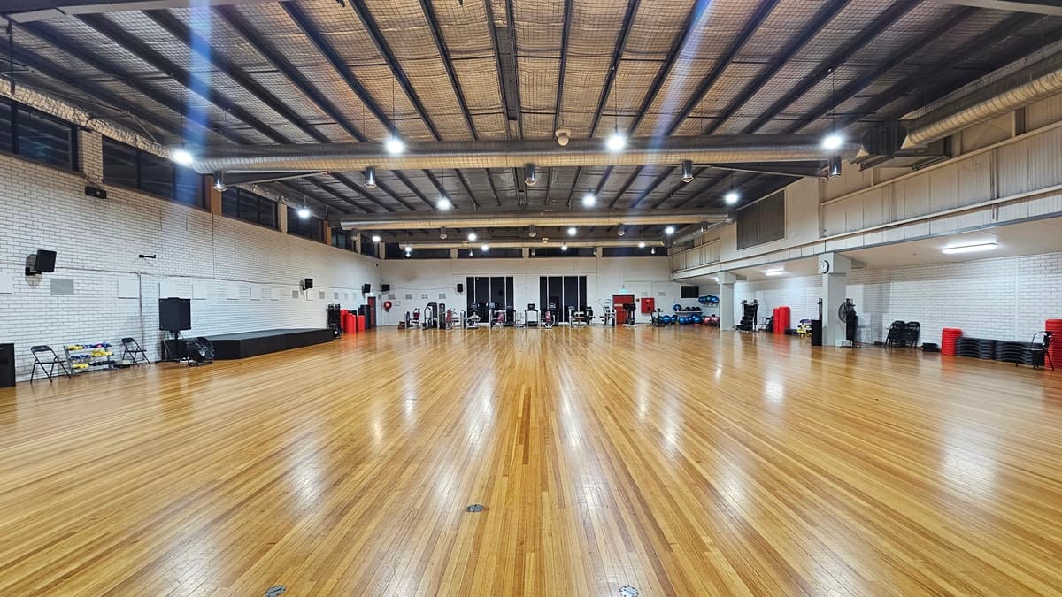 A spacious indoor gym with glossy wooden flooring and a ceiling featuring exposed steel beams and strip lighting. On the left, individuals perform exercises on mats with step platforms; on the right, stationary bikes and red lockers align the wall. Various exercise equipment are arranged on the sides.