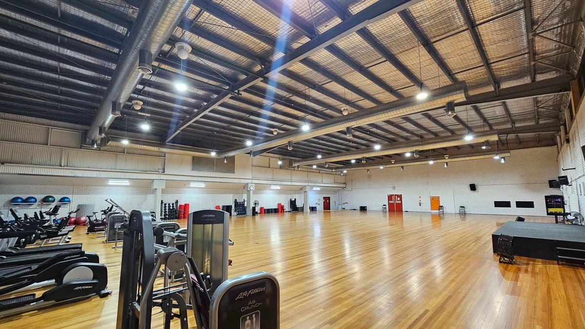 A spacious gymnasium features a high ceiling with exposed ductwork and suspended lights. On the left, cardio machines such as treadmills and ellipticals (LifeFitness brand) are positioned, while the right side displays resistance machines, stability balls, and free weights. Additionally, there is a stage equipped with speakers and fans on the right. The flooring is made of timber and the walls are constructed from white bricks.