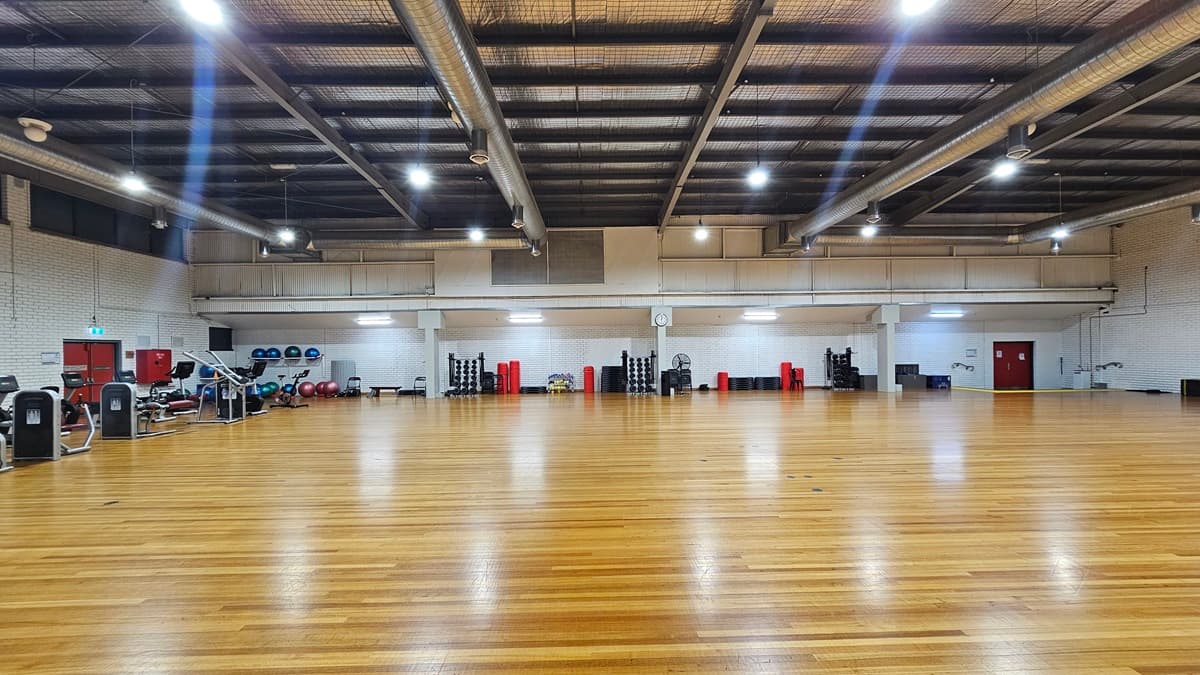 A spacious gymnasium boasts a high ceiling with exposed ductwork and suspended lights. To the left, an array of cardio machines including treadmills and ellipticals are arranged, alongside stability balls. The central area is equipped with resistance machines, more stability balls, and free weights. The floor is made of timber, complementing the white brick walls.