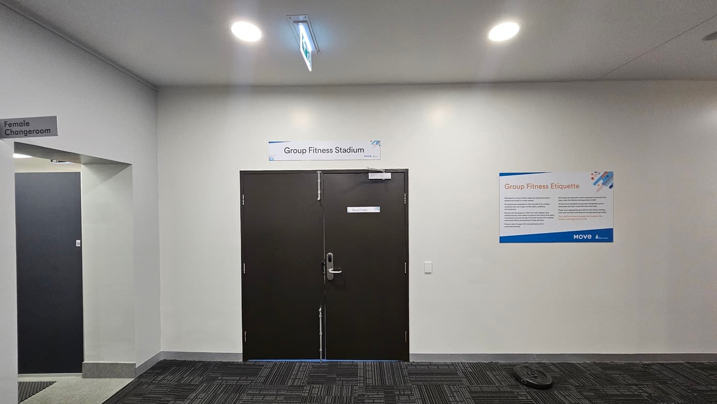 Interior of a facility with a door labelled "Group Fitness Studio" in the centre, flanked by signs. To the left, a sign for "Female Changeroom," to the right, an informational poster. Floor features a patterned dark carpet, while the ceiling has recessed lighting.