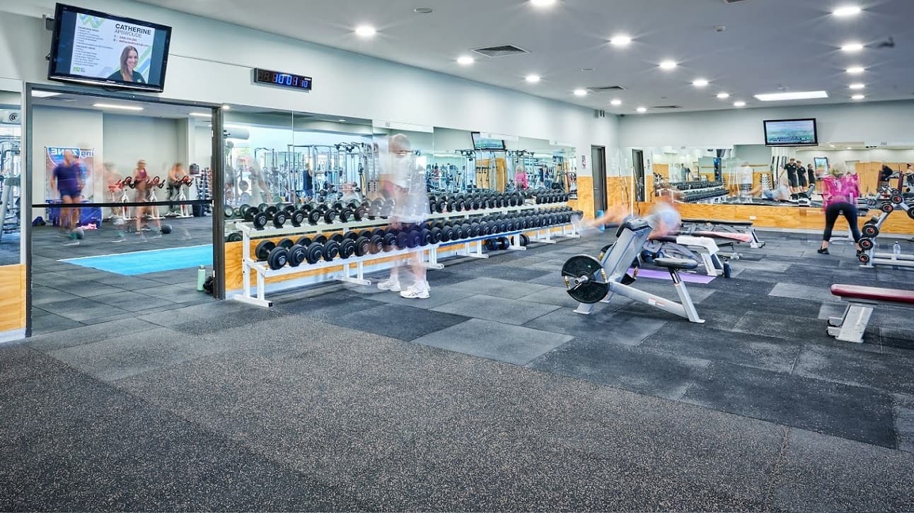 On the left, a row of dumbbells is arranged on a rack adjacent to a mirrored wall, which reflects other gym patrons. To the right, gym-goers are using exercise equipment. The background features additional fitness machinery and flat-screen TVs. In the foreground, a gym-goer is positioned on a bench press. The floor is covered with speckled rubber tiles, and the ceiling is white, equipped with recessed lighting.