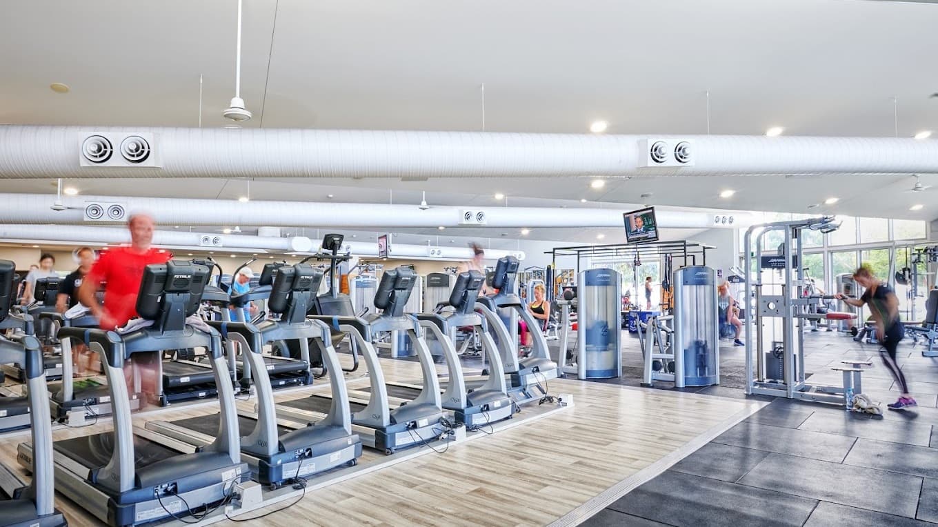 Inside the gym, a row of treadmills equipped with personal screens lines the left side. On the right, weight training equipment including a power rack and weight plates is available. The floor is made of timber, and the white ceiling features cylindrical air ducts and recessed lights. The walls are adorned with mirrors and windows, and gym-goers using the machines are visible.