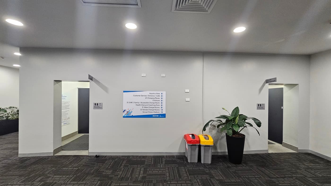 Two separate doors lead to male and female changing rooms. A large poster with directions is affixed to the white wall. Beside this, two rubbish bins are placed next to an indoor plant. The walls and ceiling are both white, the latter featuring recessed lights and air conditioning. The floor is covered with grey carpet. To the left, there is a lobby where an indoor plant is set against the wall.