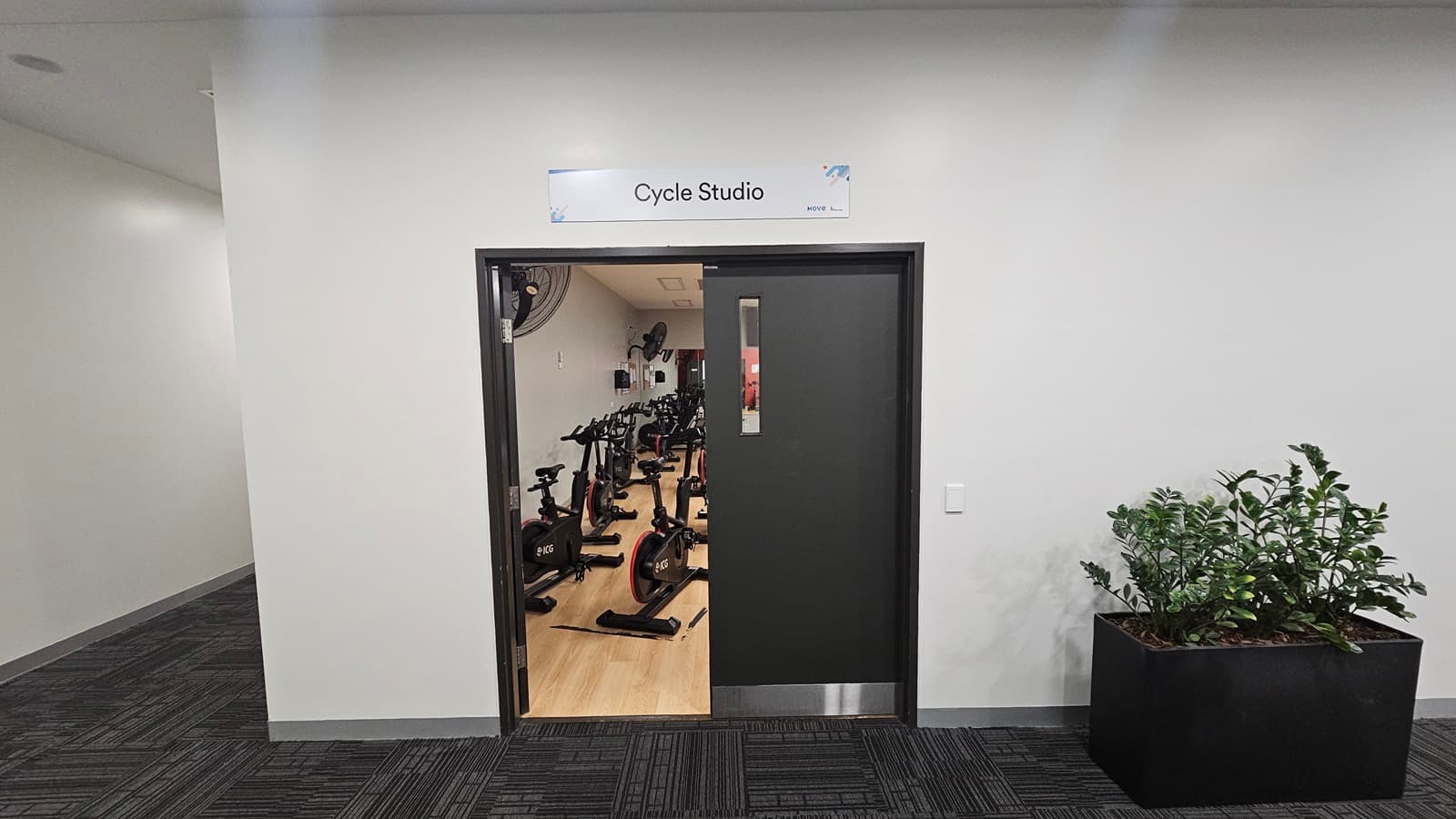 An inward-opening door leads to a Cycle Studio with timber floors and arranged stationary bikes. Above the door, a sign reads "Cycle Studio." To the right, a planter box with indoor plants is positioned. To the left, there is a lobby. The wall is painted white, and the floor is covered with grey tiles.