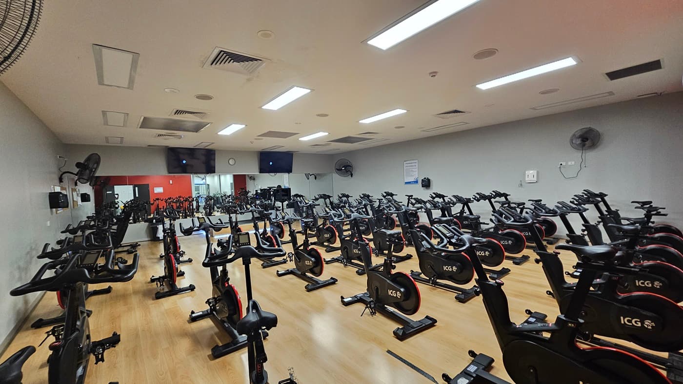 A spacious indoor cycling studio, symmetrically arranged with rows of stationary exercise bikes on both the left and right sides. Each bike is equipped with a red flywheel and a black saddle. The flooring is made of timber and the middle wall is painted red, featuring a black door and a section covered with mirrors. The left wall has light colours with wall fans affixed.Flatscreen TVs are affixed to the upper walls.