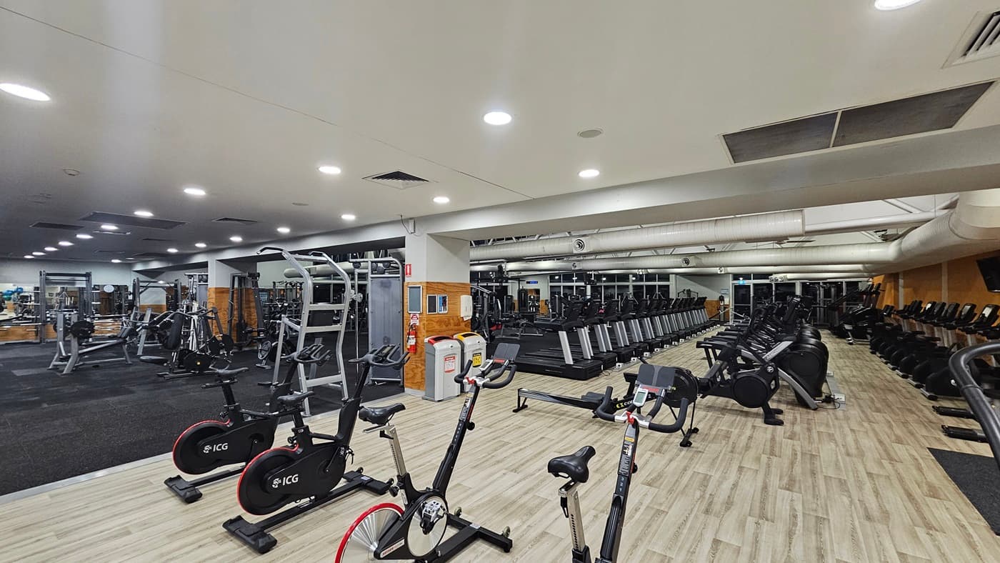 The interior of the gym includes a variety of equipment. On the left, there is a row of treadmills with a free weights area situated further back. The right side features stationary bikes in the foreground and elliptical machines positioned behind them. Timber flooring spans the area on the right and black rubber tiles fills the left. The white ceiling is equipped with aircon vents and recessed lights. Rubbish bins are strategically placed throughout the space.