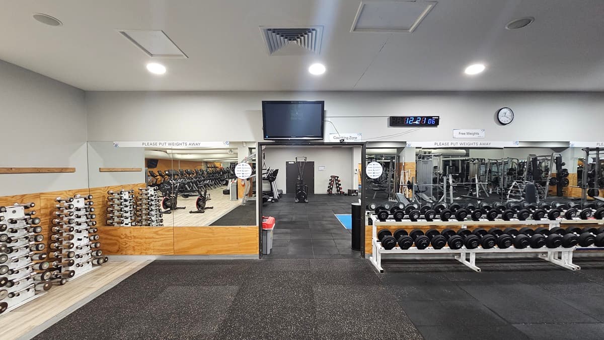 On the left, a rack of dumbbells in ascending weights is aligned next to a wall clad partly with mirrors. In the centre, a mounted television hangs above a digital clock displaying the time. To the right, additional dumbbell racks and weight training machines, including cable systems and leg press machines, are visible. Reflective mirrors span the right-hand side. The floor is covered with heavy-duty, interlocking rubber tiles, and the ceiling hosts recessed lighting fixtures.