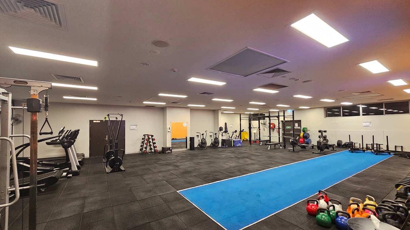Fitness machines are arranged on the left, including treadmills, elliptical trainers, and Smith Machines. On the right, free weights, kettlebells, and a plyometric box are arranged. The central area features a long blue sprint mat. The ceiling is white with aircon vents, fluorescent lighting. The floor is covered with black rubber tiles, and mirrors are fitted on the far wall.
