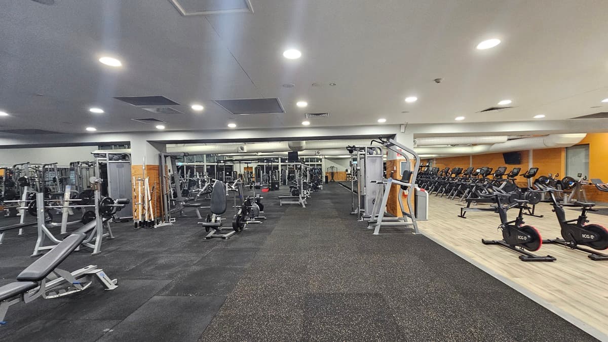 The interior of the gym features a variety of fitness equipment. To the left, there are weight benches, barbell racks, and sets of dumbbells. On the right, treadmills and stationary bikes are positioned. In the centre of the room, cable machines, lat pull-down machines, and bench machines are arranged. The entire floor is covered with grey padded flooring, and above, a white paneled ceiling is fitted with recessed lights.