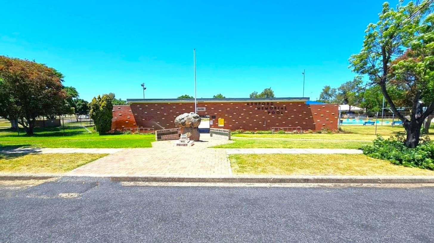 A brick building with the words "Ashford Memorial Pool" is in the centre background. In front, a stone monument with "Lest We Forget" is surrounded by a paved area. To the left and right, there are trees and grass. A swimming pool with people is visible on the right, enclosed by a fence. The foreground shows a paved road and a grassy verge.
