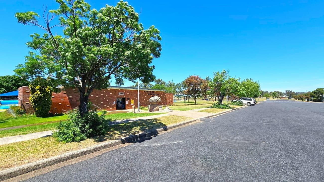 A street scene with a brick building on the left, partially obscured by a large tree. The building has a sign above the entrance and a small path leading to it. A swimming pool with a blue shade structure is visible behind the building. On the right, a paved road runs alongside a grassy area with more trees and a parked white vehicle. The foreground features a kerb and a grassy verge.