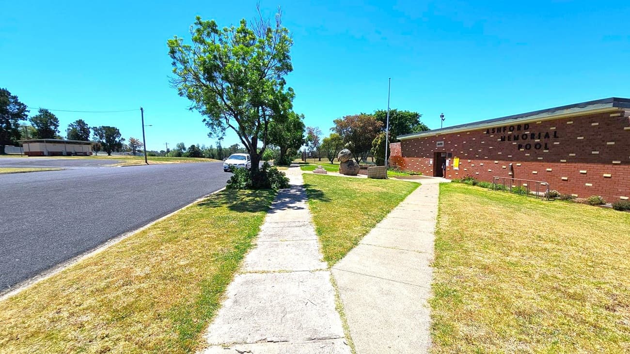 A paved footpath runs through the centre, flanked by grass on both sides. On the right, a brick building with the words "ASHFORD MEMORIAL POOL" is visible. A concrete path leads to its entrance. On the left, a tree provides shade over a parked white car on the street. In the background, more trees and a small building are visible across the road. The sky is clear and blue.