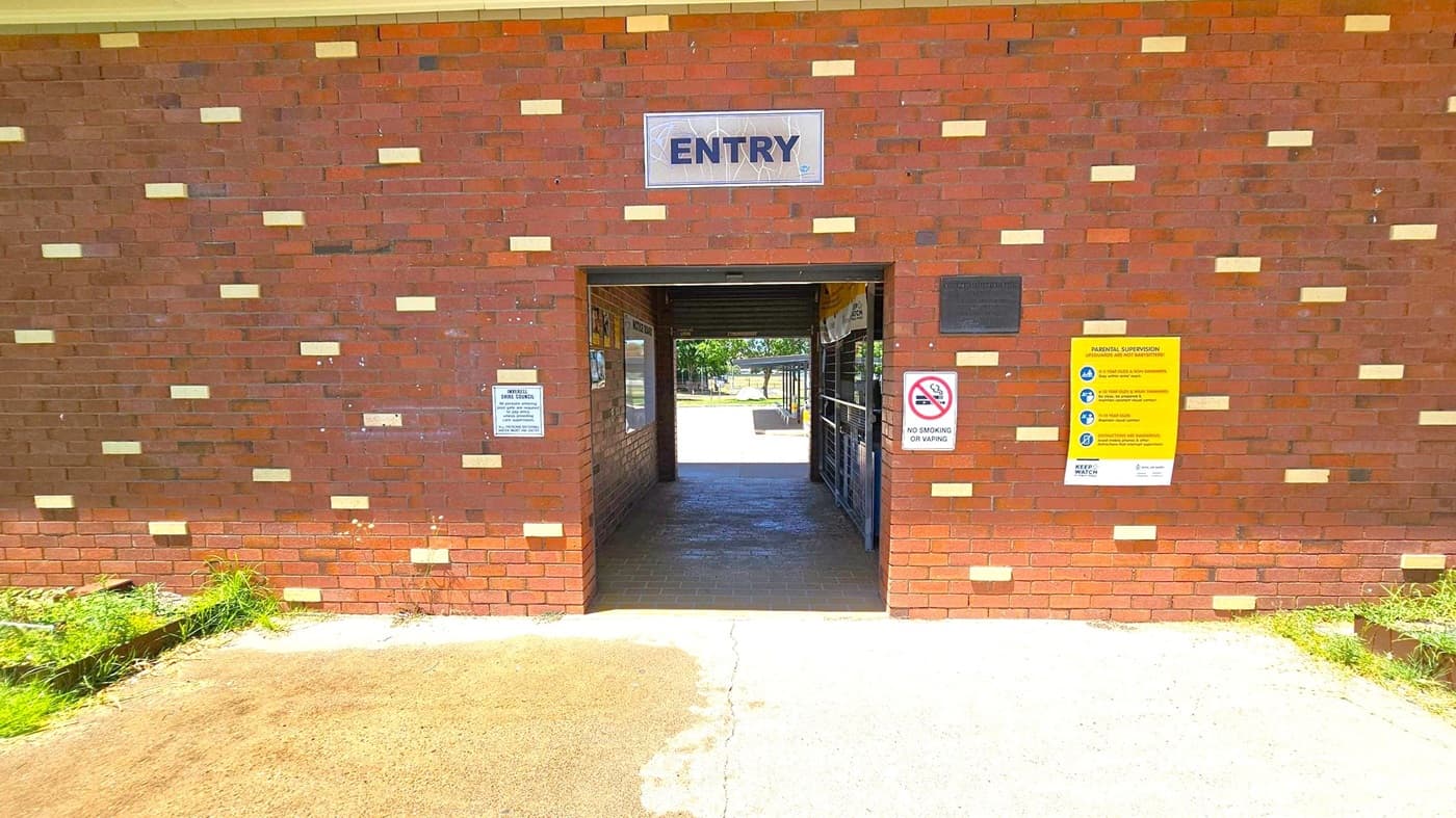 A brick wall with a rectangular entryway in the centre. Above the entry, a sign reads "ENTRY". On the right side of the entry, there are two signs: one prohibiting smoking or vaping, and another with parental supervision guidelines. On the left, a smaller sign is visible. The ground is a mix of concrete and grass, with some greenery on the left. Through the entryway, a glimpse of an outdoor area with trees and a pathway is visible.