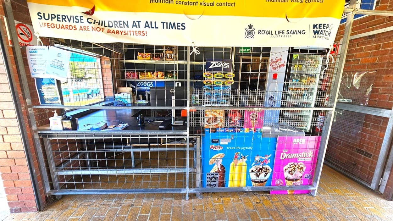 A kiosk with a metal grid barrier in front. On the left, a brick wall with signs and a hand sanitiser dispenser on a counter. In the middle, a window displaying snacks and a computer monitor. On the right, a refrigerator with drinks and ice cream advertisements, including Peters and Drumstick. A large banner above reads "SUPERVISE CHILDREN AT ALL TIMES" with logos for Royal Life Saving Australia and Keep Watch. The floor is tiled with a yellow-brown pattern.