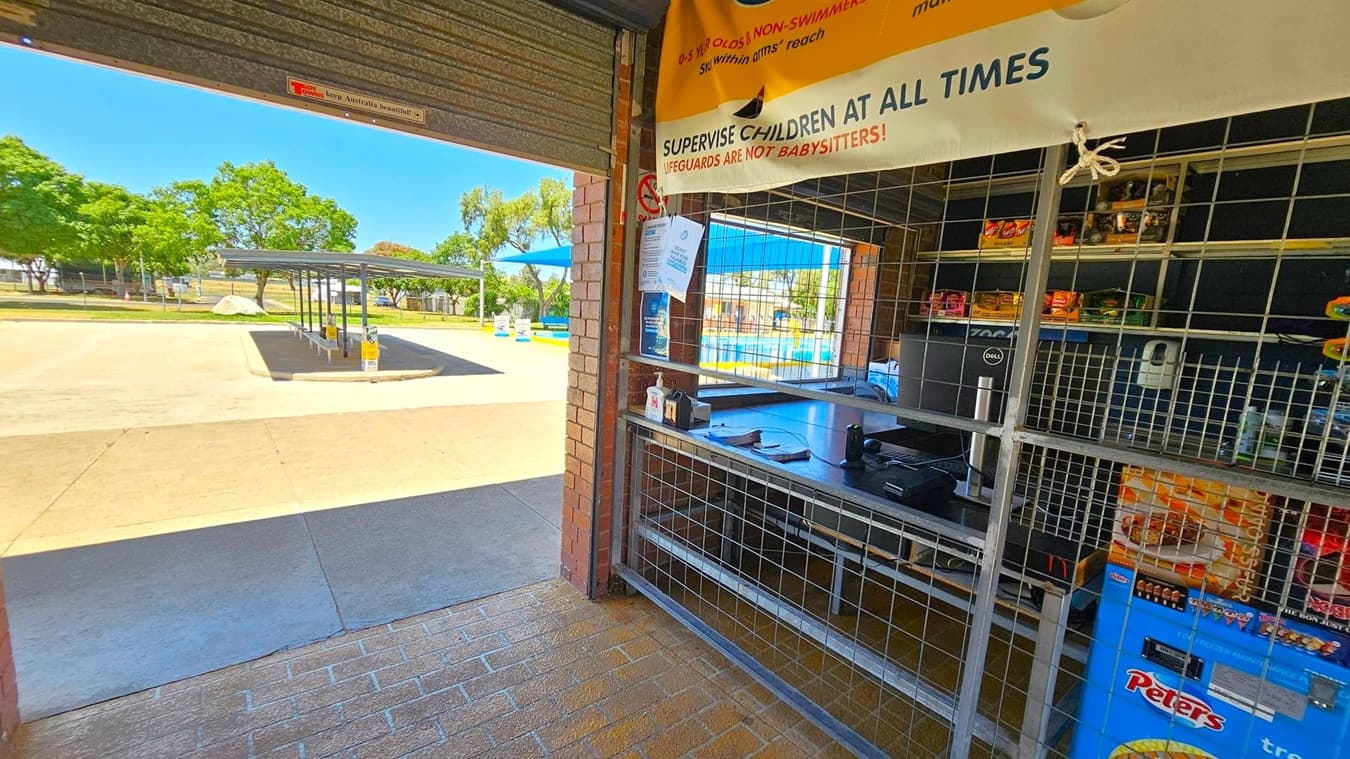 On the left, an open area with a paved surface and a covered seating area. Trees and a grassy area are visible in the background. On the right, a kiosk with a metal grid, displaying snacks and a freezer with ice cream branding. A sign above advises supervising children. The floor is tiled, and the ceiling is not visible.