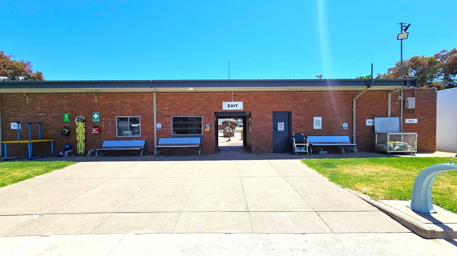 A brick building with a flat roof and an "EXIT" sign above a central doorway. On either side of the door are benches and various safety equipment, including a yellow stretcher and first aid signs. To the right, a door marked "MALE" is visible. The foreground features a paved area with grass on either side. A metal structure is on the right edge. Trees are visible in the background.