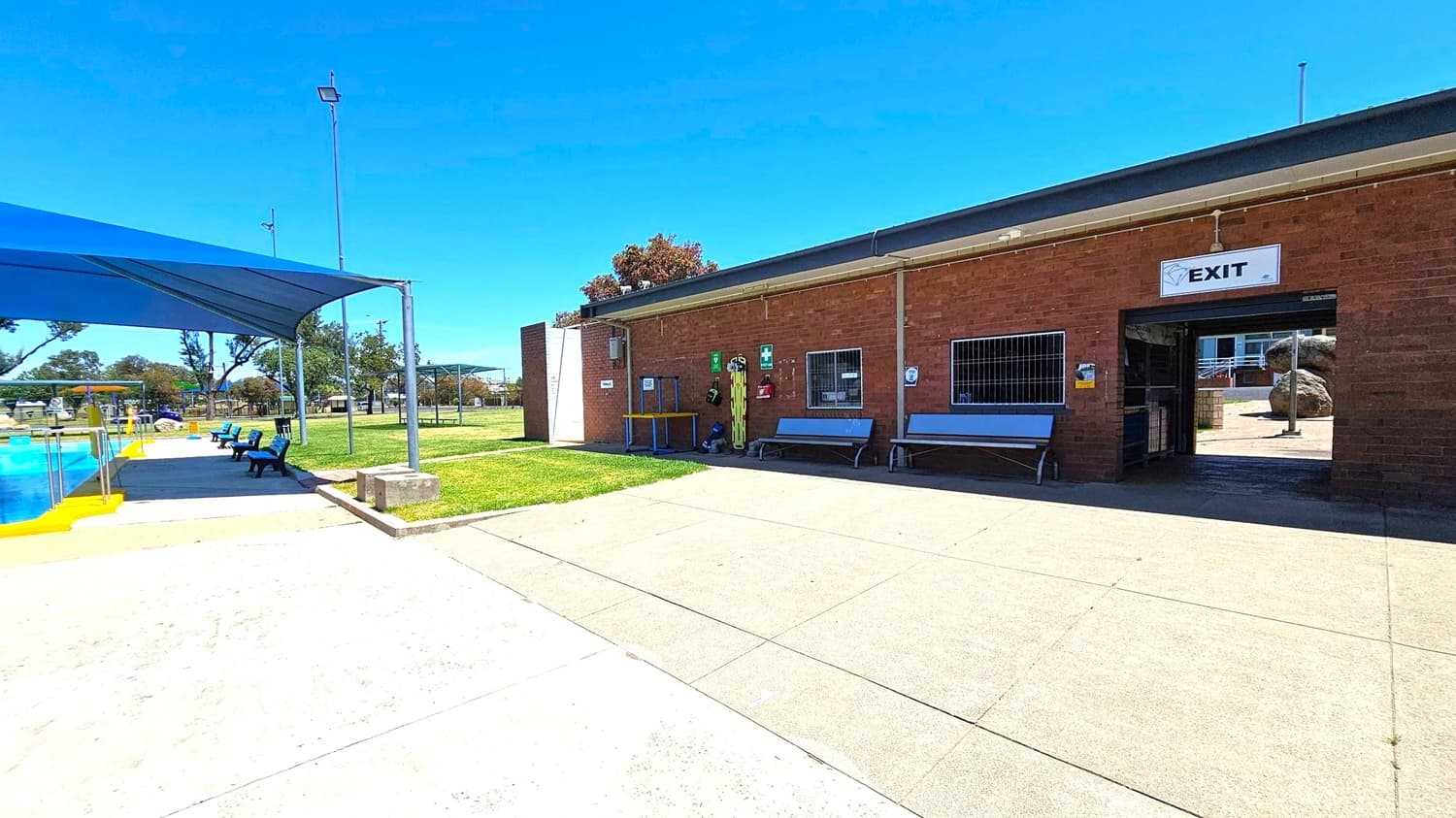 A swimming pool area with a blue shade structure on the left, covering benches and a pool with yellow edges. The ground is a mix of concrete and grass. On the right, a brick building with an "EXIT" sign above a doorway. Benches and safety equipment are against the building's wall. In the background, trees and more shaded areas are visible. The sky is clear and blue.