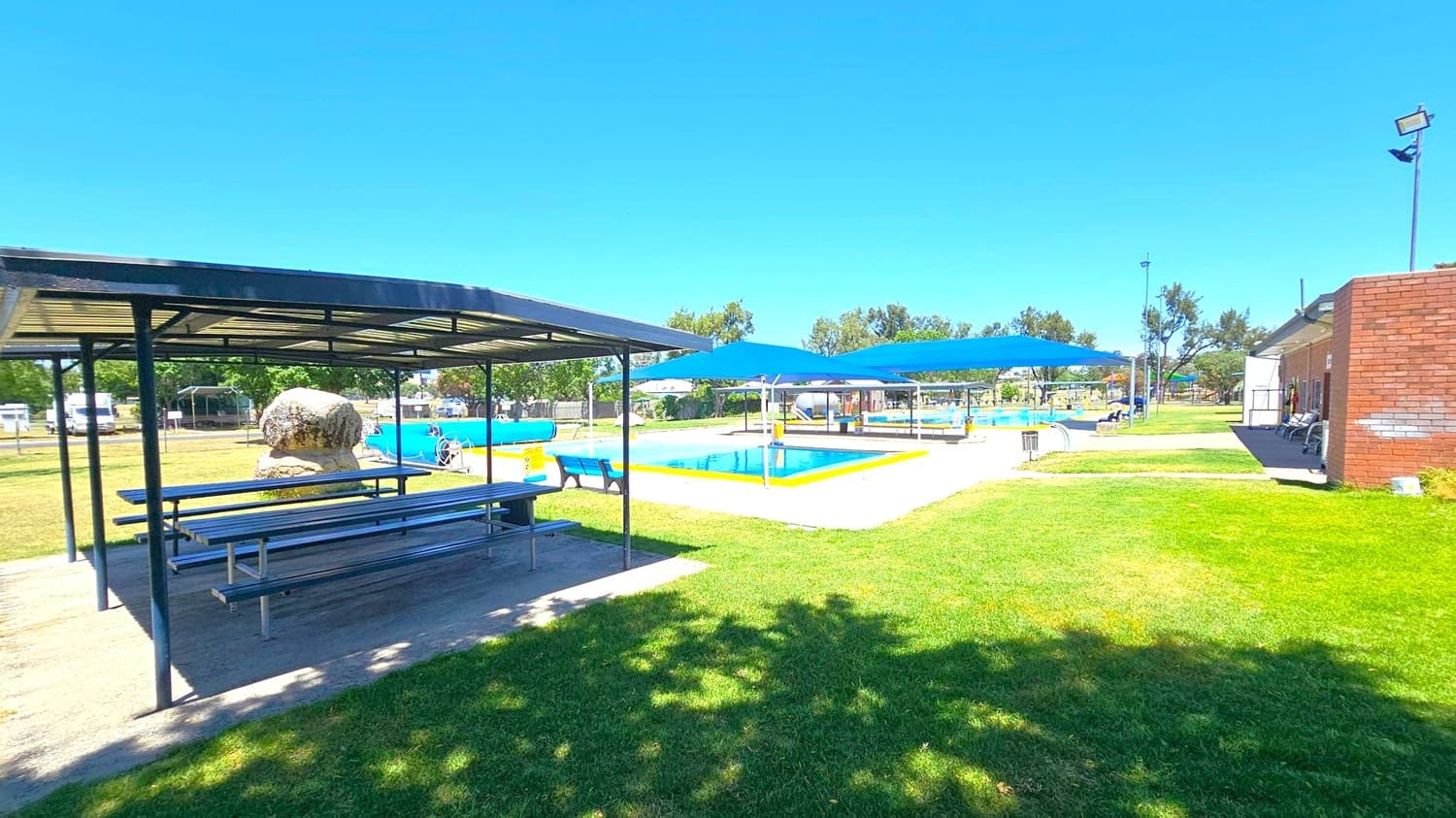 A recreational area with a covered picnic table on the left, featuring metal benches and a roof. In the centre, there are two swimming pools with blue shade sails above them. The pools are surrounded by a sandy area and green grass. On the right, a brick building with outdoor seating is visible. Trees and a fence line the background, and the sky is clear.