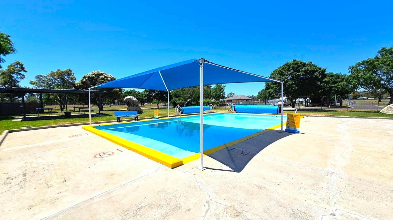 A small rectangular swimming pool with bright yellow edges is in the centre, covered by a blue shade sail supported by white poles. The pool is surrounded by a light-coloured concrete surface with some visible cracks. On the left, there is a blue bench and a large rock. In the background, there are several trees and a fenced area. A sign with safety instructions is placed near the pool.