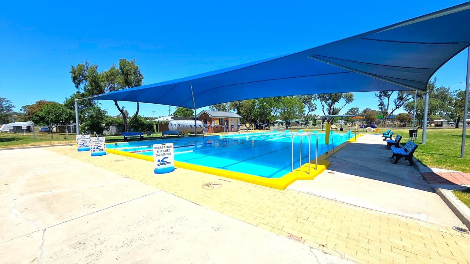 A swimming pool with bright blue water is covered by a large blue shade sail. The pool has yellow edges and metal handrails. Signs around the pool indicate "Recreation & Leisure Swimming." On the right, there are several blue benches on a paved area. In the background, there are trees, a small building, and a grassy area. The foreground features a mix of concrete and brick paving.