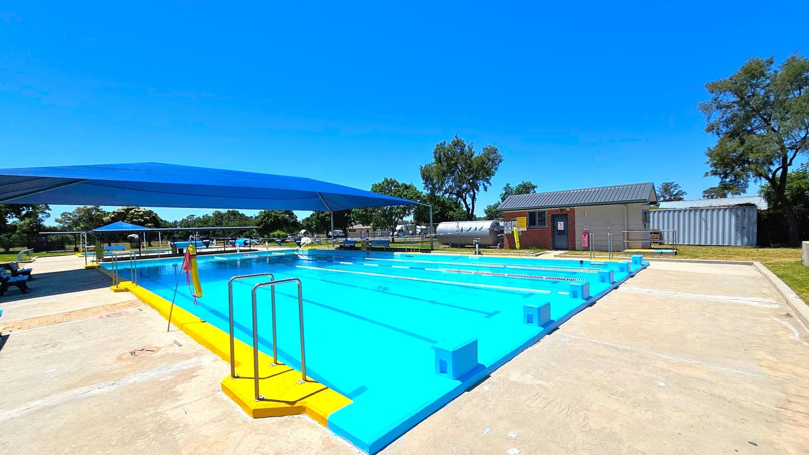 A rectangular swimming pool with bright blue water is in the centre, surrounded by a yellow border. On the left, a blue shade structure covers part of the pool area. There are metal ladders for entry into the pool. In the background, a small building with a grey roof and a large cylindrical tank are visible. Trees and grassy areas surround the pool, and the ground is a mix of concrete and grass.