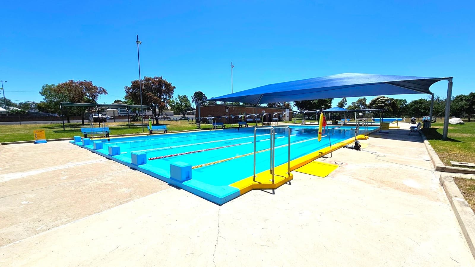 A rectangular outdoor swimming pool with bright blue water and yellow edges. Lanes are marked with red and yellow ropes. A blue shade structure covers part of the pool. Metal ladders are positioned at the pool's edge. Surrounding the pool is a concrete deck with some grass areas visible. Benches and a few trees are in the background, along with a fence and some buildings. A tall light pole is also visible.