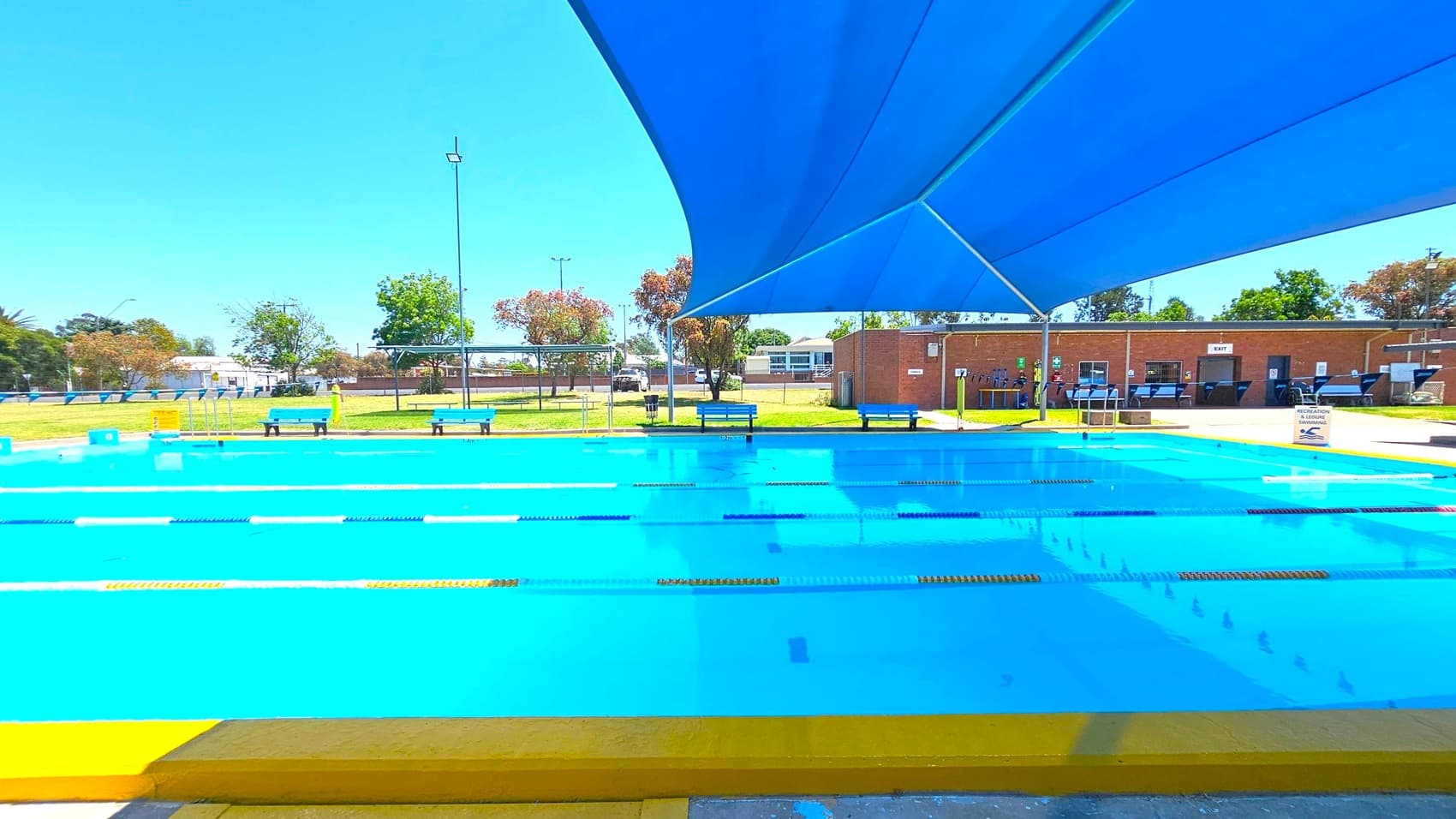An outdoor swimming pool with clear blue water, divided into lanes by floating ropes. A large blue shade cloth covers part of the pool. Several blue benches are positioned around the pool's perimeter. In the background, there is a grassy area with trees and a brick building with an "EXIT" sign. The sky is clear and bright.