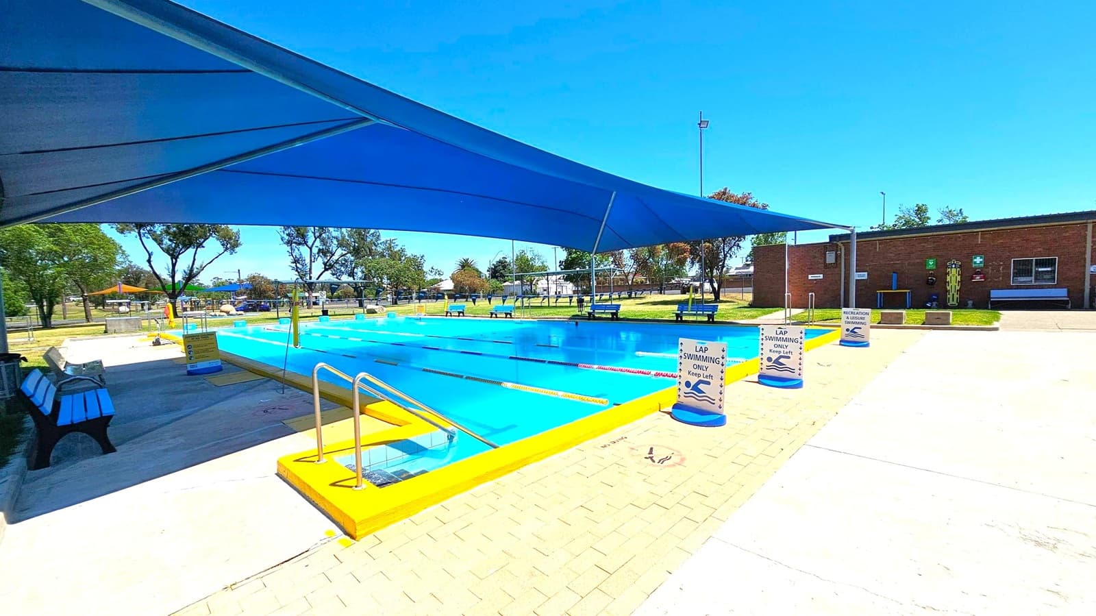 A swimming pool with clear blue water is in the centre, surrounded by a yellow border. A large blue shade structure covers part of the pool. Signs around the pool indicate "Lap Swimming Only." On the left, there are benches and trees in the background. On the right, a brick building with windows and a grassy area is visible. The ground is a mix of concrete and brick paving.