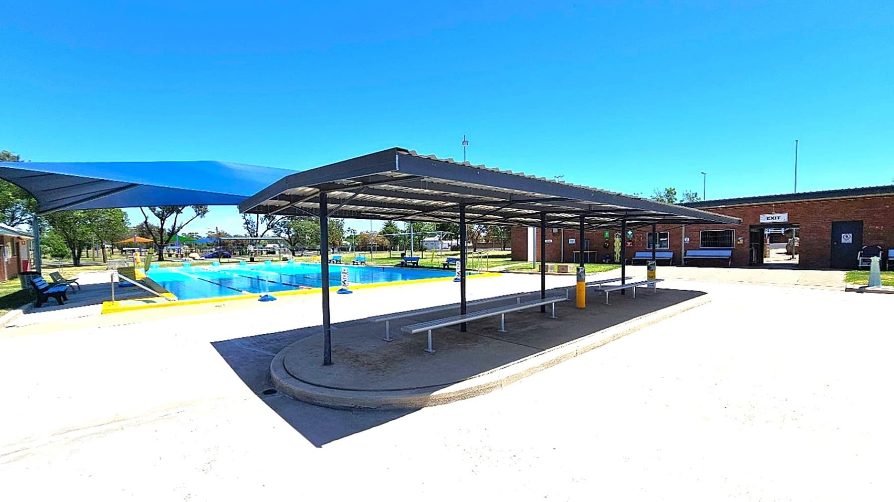 A swimming pool area with a blue shade structure on the left, covering part of the pool. In the centre, there's a covered seating area with benches underneath a metal roof. The pool is surrounded by a yellow border, and there are trees and benches in the background. On the right, a brick building with an "EXIT" sign above a doorway. The ground is paved with light-coloured concrete.