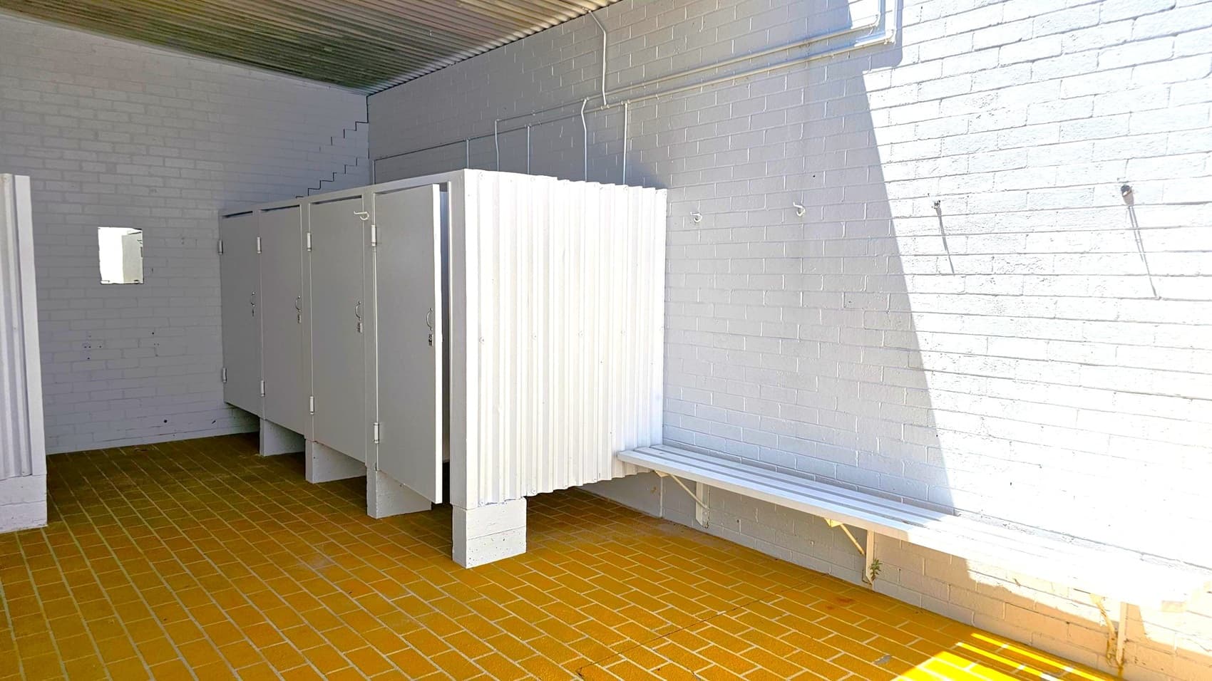 A changing room with white brick walls and a corrugated metal ceiling. On the left, there are several closed cubicles with white doors. On the right, a long white bench is attached to the wall, with hooks above it. The floor is covered with brown tiles. A small window is visible on the left wall.