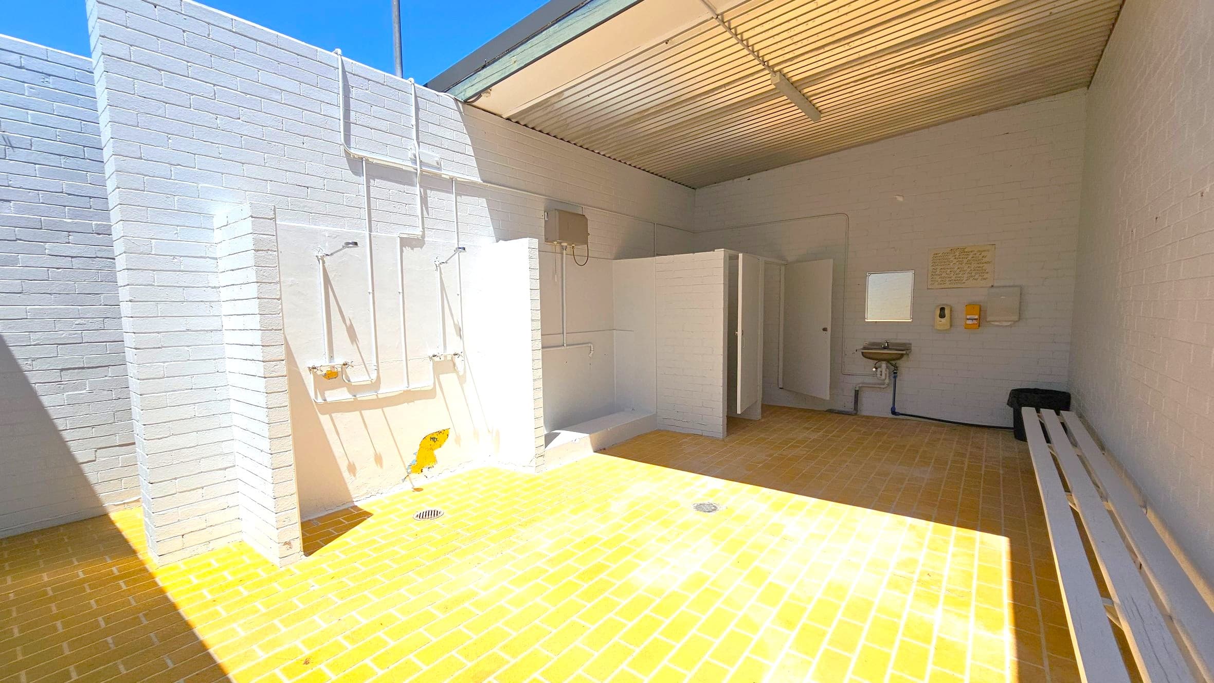 An outdoor shower area with white brick walls and a yellow brick floor. On the left, there are two shower heads with exposed piping. In the centre, there are open cubicles with partitions. On the right, a sink is mounted on the wall with a mirror above it, and a bench is positioned against the wall. The ceiling is made of corrugated metal with a fluorescent light fixture.