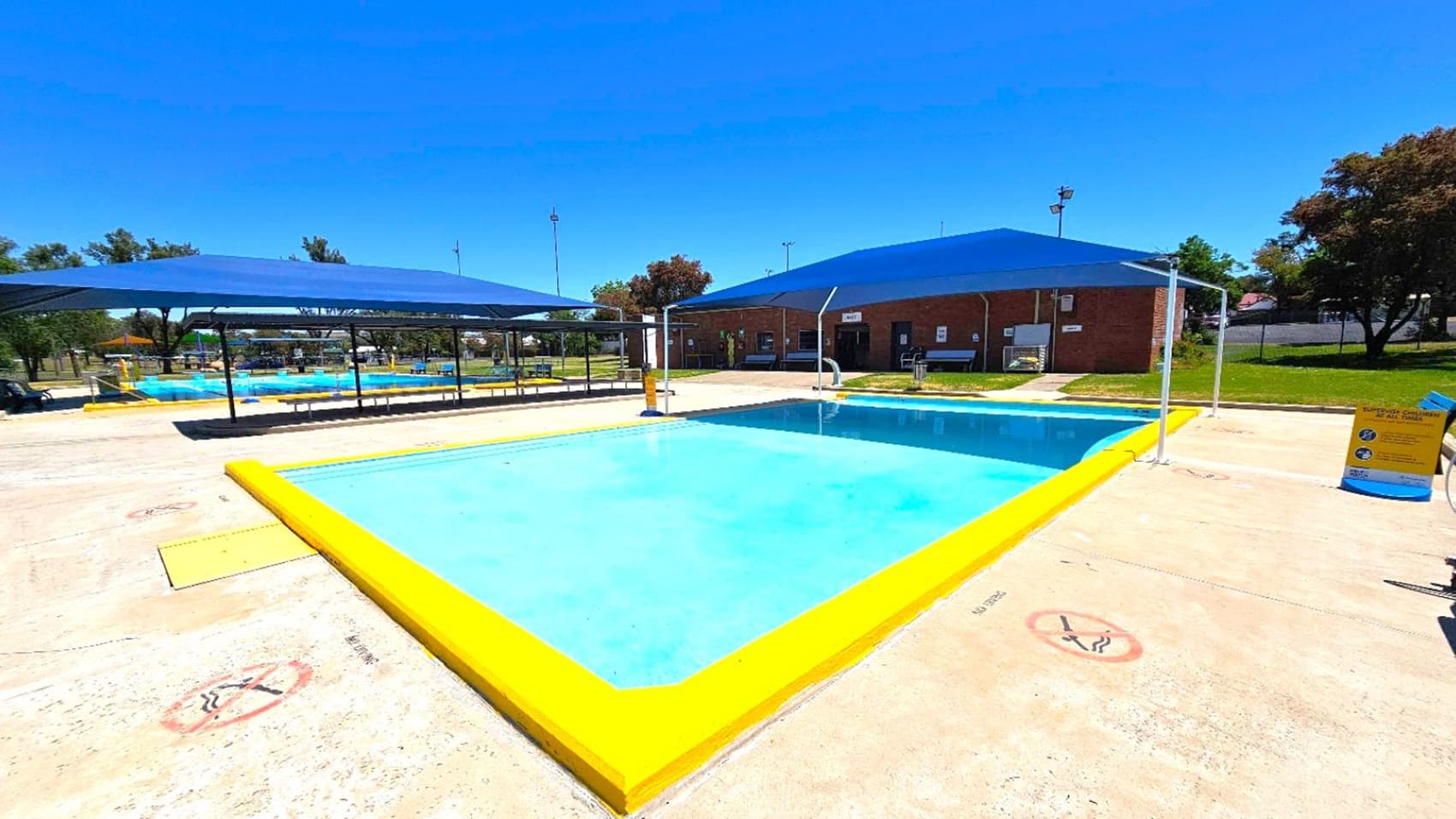 A swimming pool with a bright yellow border is in the foreground, surrounded by a concrete deck with "No Diving" signs. Two blue shade structures cover seating areas on either side of the pool. In the background, a brick building with doors and windows is visible, along with grassy areas and trees. A sign with pool rules is on the right side near the pool. The sky is clear and blue.