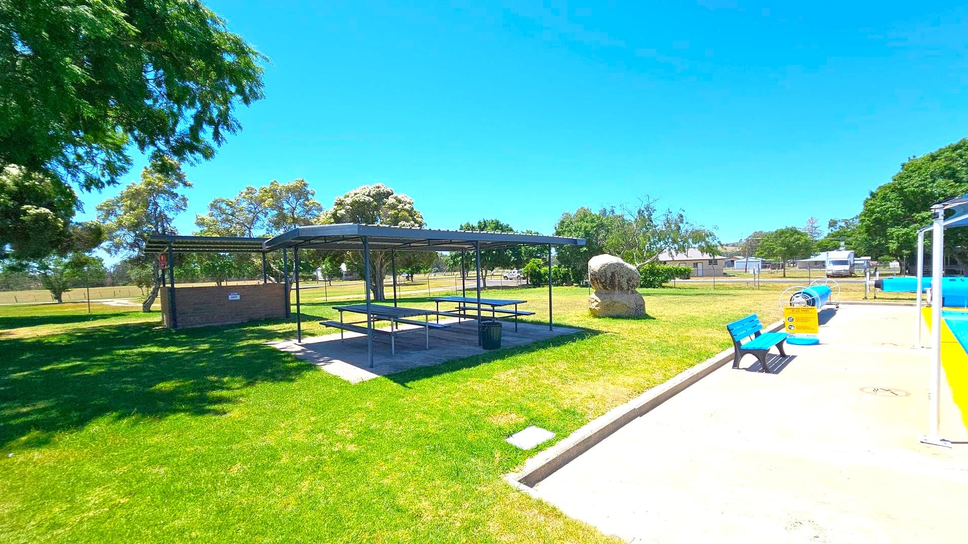 A park scene with a covered picnic area in the centre, featuring several tables and benches on a concrete slab. To the right, a blue bench sits on a paved area next to a yellow and blue play structure. A large rock is visible on the grass nearby. Trees with dense foliage are scattered throughout the background, and a fence runs along the perimeter. The grass is well-maintained, and the sky is clear.