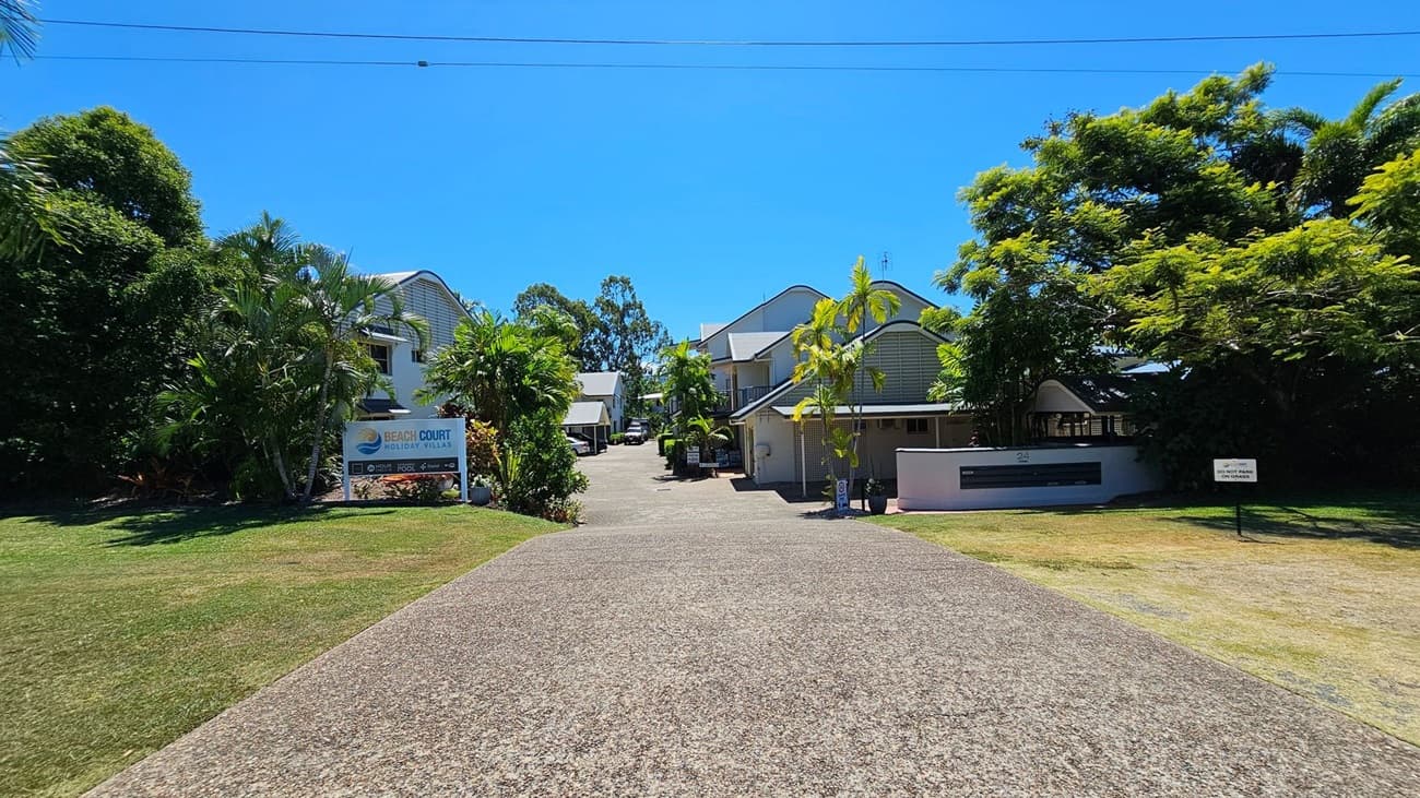 A paved driveway stretches from the foreground to the middle, flanked by green grass on both sides. To the left, a signboard for "Beach Court Holiday Villas" is set amidst dense palm trees. To the right, a white building with a dark sign is partially visible behind smaller trees. In the middle, modern two-storey buildings with light facades and peaked roofs line the driveway, with additional trees and greenery between them.