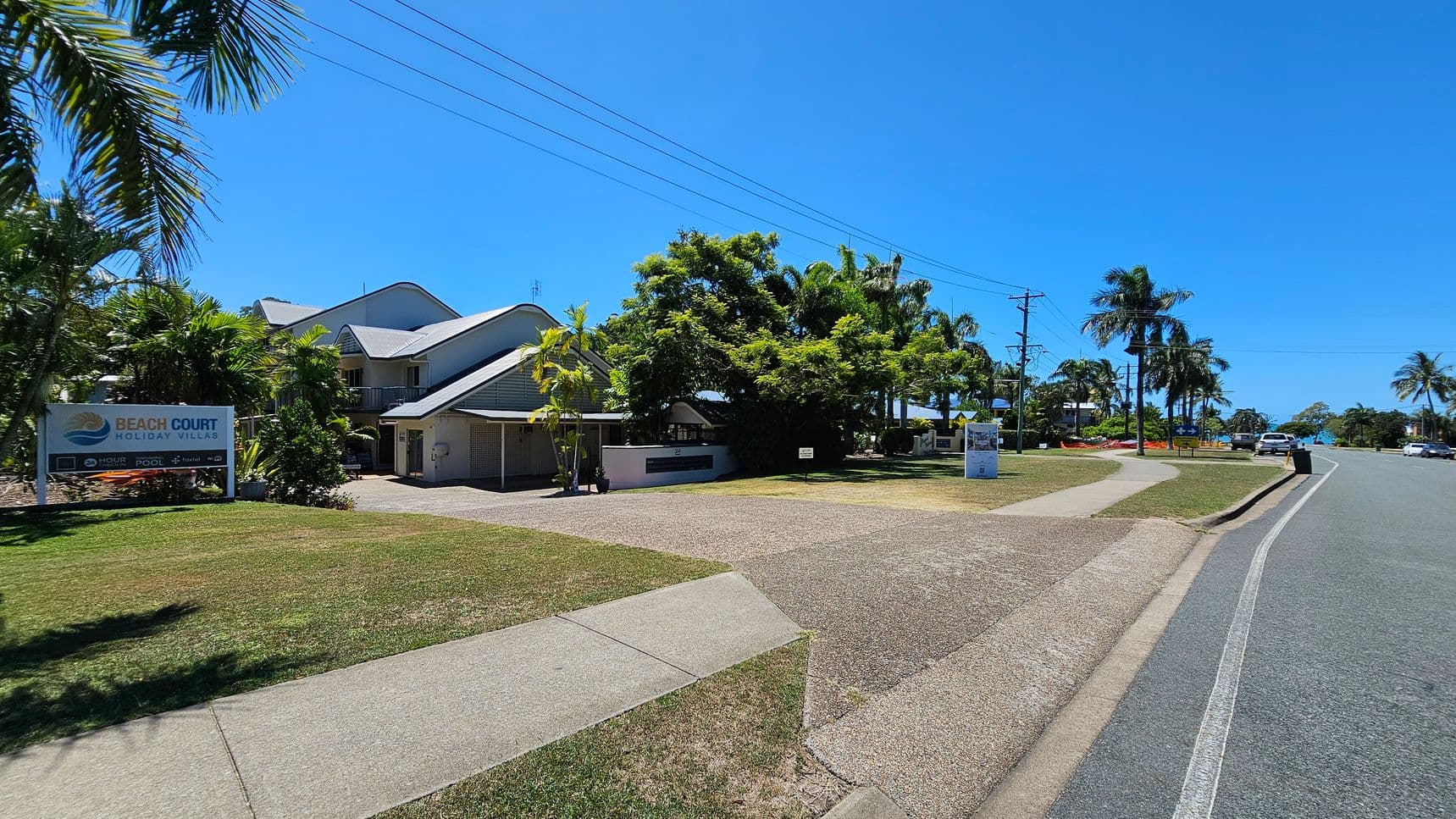 On the left, there is a two-storey building with a grey roof surrounded by palm trees and greenery. A signboard reading “Beach Court Holiday Villas” stands on the grass. On the right, there is a paved road bordered by more palm trees, parked cars, and a footpath. In the middle, a driveway on a pebbled surface leads to the building. The background features more trees and low-lying structures. The foreground contains grassy patches and concrete footpaths.