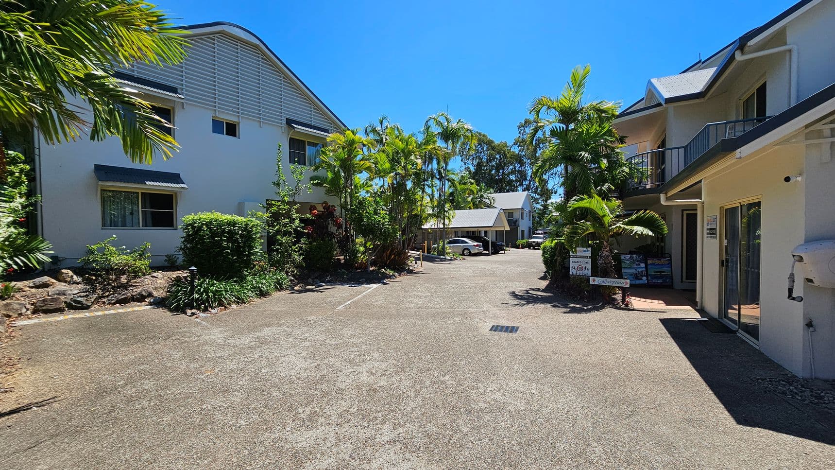 On the left, there is a white two-storey building with small awnings over the windows and greenery including bushes and palm trees. On the right, there is a similar building with balconies, glass doors, and potted plants near the entrance. In the middle, a paved area extends towards a smaller building and parked cars. The background includes more trees and shrubs. The foreground features a concrete surface with a drainage grate.
