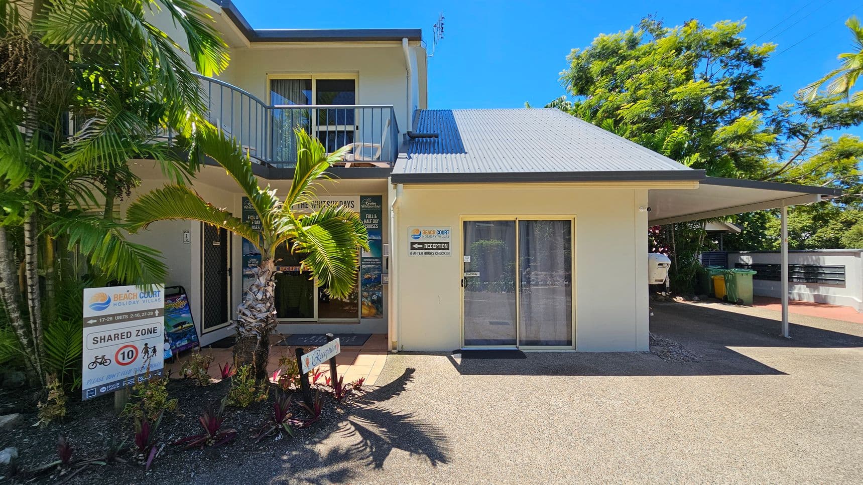 On the left, there is a two-storey cream-coloured building with a balcony, sliding glass doors, and a small palm tree in front. Below, a sign displays “Beach Court Holiday Villas” and shared zone instructions near garden beds with small plants. On the right, a single-storey section features a sliding glass door with a curtain and a covered carport. The foreground consists of a textured, pebbled surface, while the background includes trees and greenery.