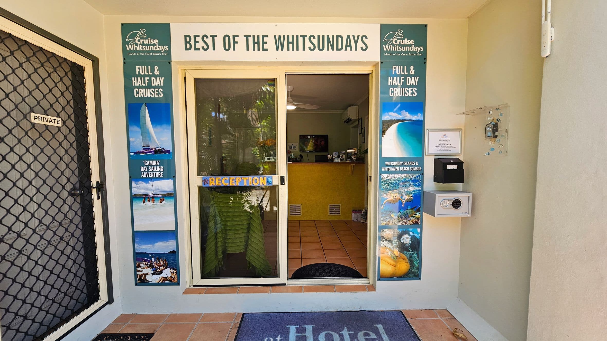 On the left, there is a black mesh door labelled "Private" beside a vertical poster advertising cruises, with images of sailing and beaches. On the right, a wall-mounted safe, noticeboard, and another vertical cruise poster showing beach scenes and marine life are visible. In the middle, glass sliding doors with a "Reception" sign lead to a tiled room with a yellow desk, and ceiling fans. The floor is terracotta tiles, with a dark mat at the entrance. The ceiling inside is white and flat.