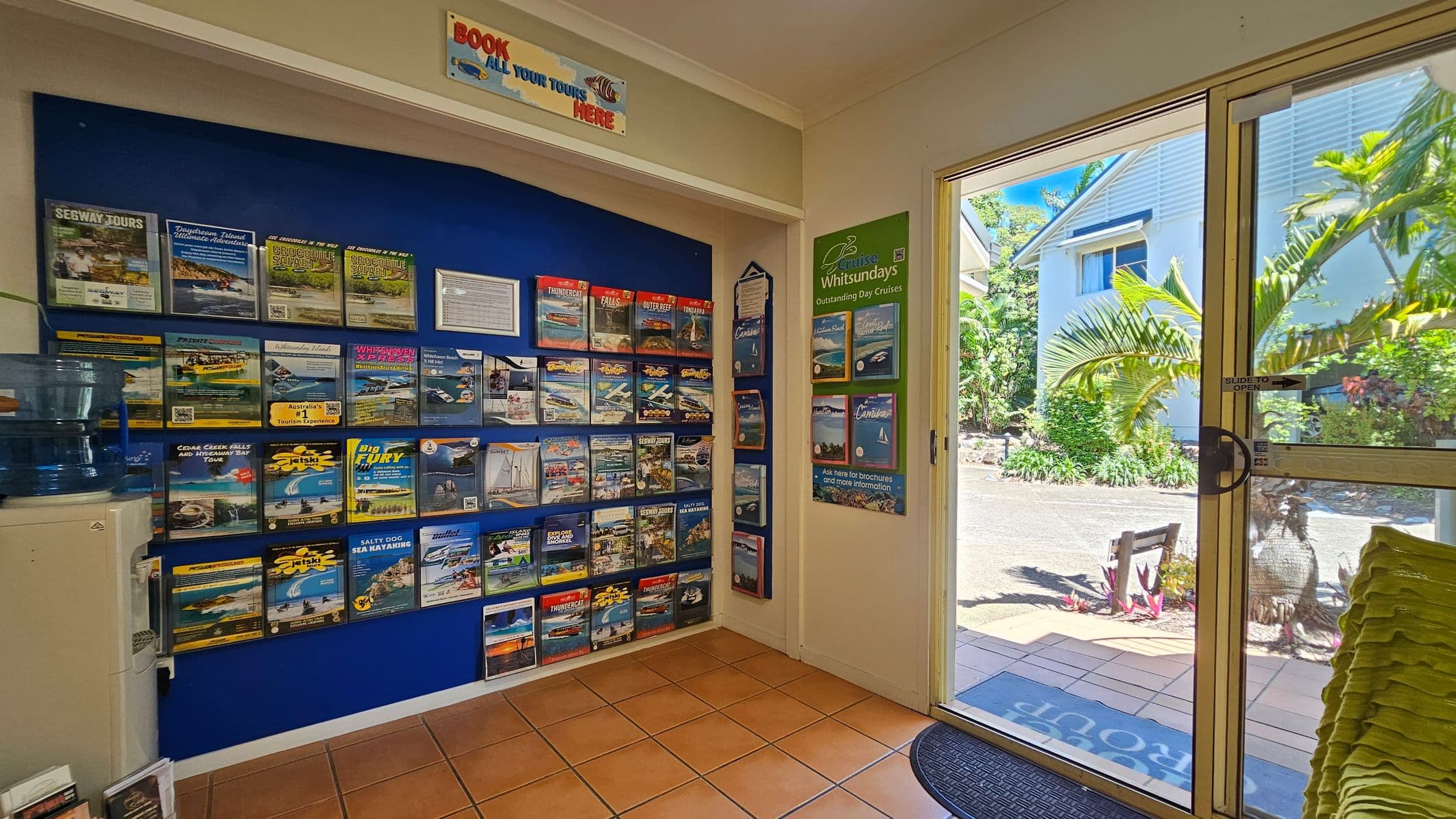 On the left, a blue wall is covered with neatly arranged brochures advertising tours and activities, with a water cooler positioned beneath them. In the middle, a sign above reads "Book all your tours here" in bold text. On the right, glass sliding doors open to an outdoor area with tropical plants, a paved path, and a white building visible in the background. The floor is tiled with terracotta-coloured squares, and the ceiling is white and flat.