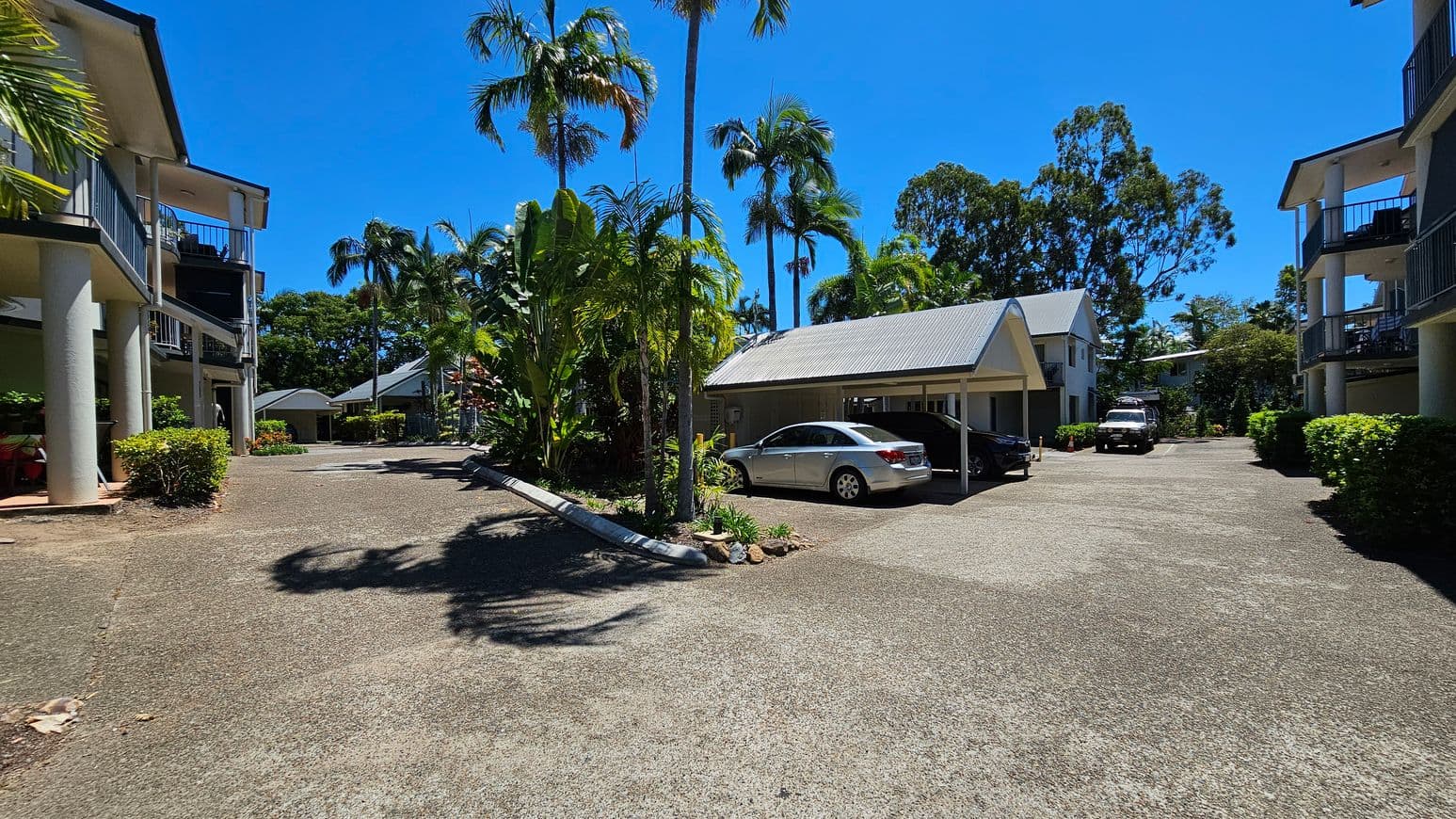 A residential area with two-storey buildings on the left and right, featuring balconies and supported by columns. In the centre, a driveway leads to a carport with a silver car parked underneath. Lush greenery, including palm trees and shrubs, surrounds the area. The ground is paved, and the sky is clear.