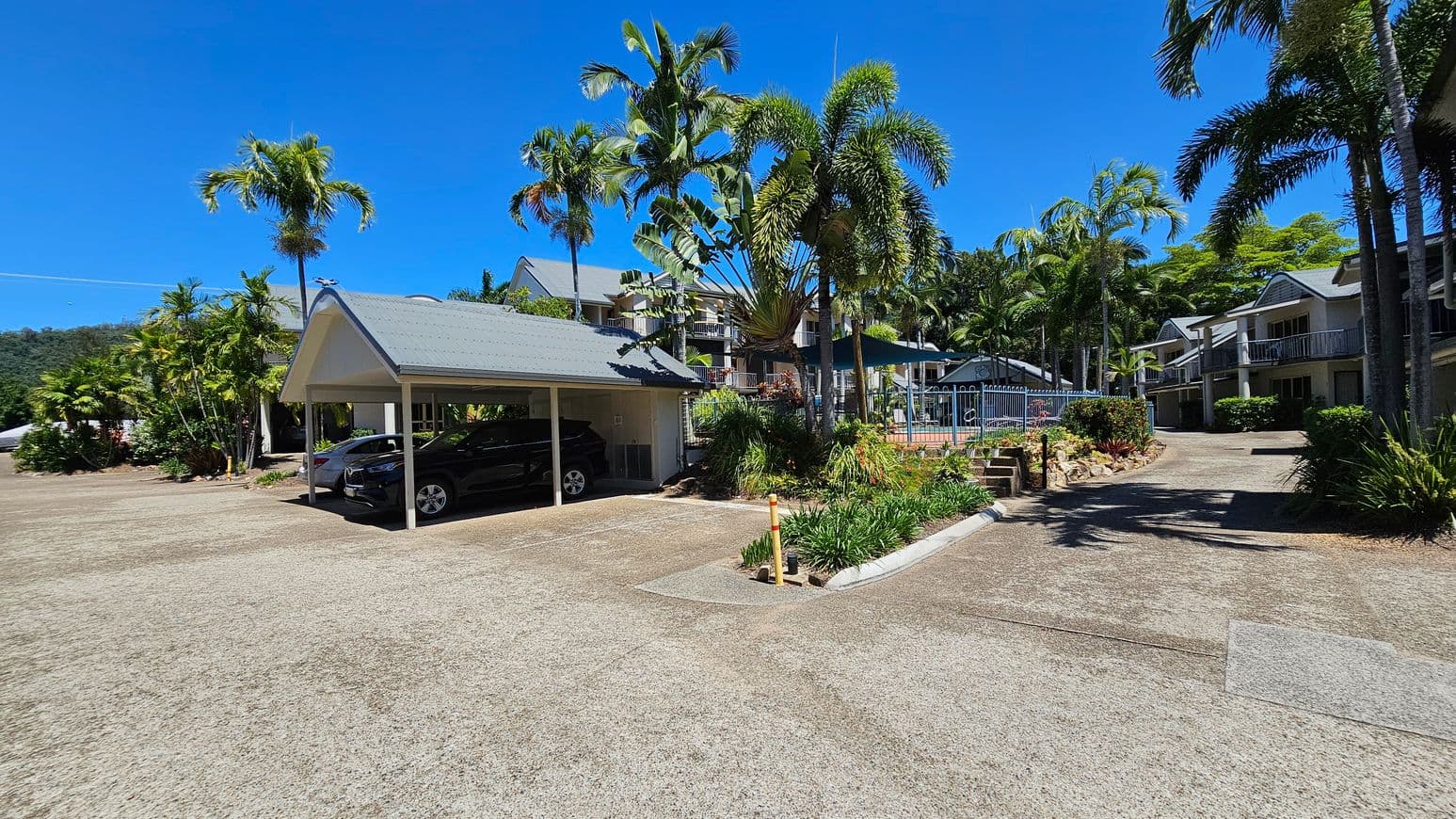 A carport on the left shelters two cars, surrounded by lush palm trees and greenery. In the centre, a pathway leads to a fenced pool area with more tropical plants. On the right, there are two-storey buildings with balconies, also surrounded by palm trees and shrubs. The ground is a mix of concrete and patches of grass.